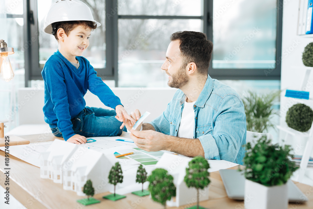 Advice needed. Charming young architect showing a color chart to his little son in a white hard hat and asking him to choose a color Advice needed. Charming young architect showing a color chart to his little son in a white hard hat and asking him to choose a color