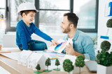 I need advice. Pleasant bristled architect sitting next to his son and showing him a color chart, asking for advice, while the boy pointing at the color of his choice