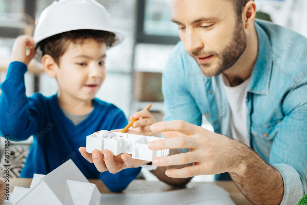 Thorough examination. Handsome young father and his little son in a white hard hat sitting at the table and examining a 3D house layout model Thorough examination. Handsome young father and his little son in a white hard hat sitting at the table and examining a 3D house layout model