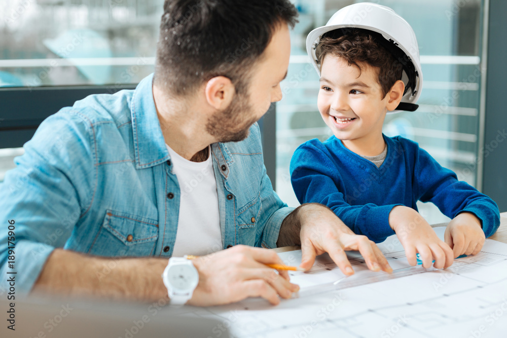 Best help. Adorable little boy sitting at the table next to his father and helping him to draw a blueprint while exchanging smiles with the man Best help. Adorable little boy sitting at the table next to his father and helping him to draw a blueprint while exchanging smiles with the man