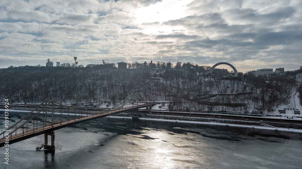 Aerial top view of pedestrian Park bridge in winter and Dnieper river from above, snow Kyiv cityscape, city of Kiev skyline, Ukraine
