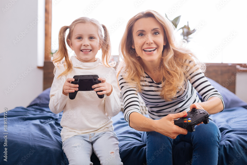 Playing with mum. Adorable content fair-haired girl laughing and holding a remote control playing with her mom while sitting on the bed Playing with mum. Adorable content fair-haired girl laughing and holding a remote control playing with her mom while sitting on the bed