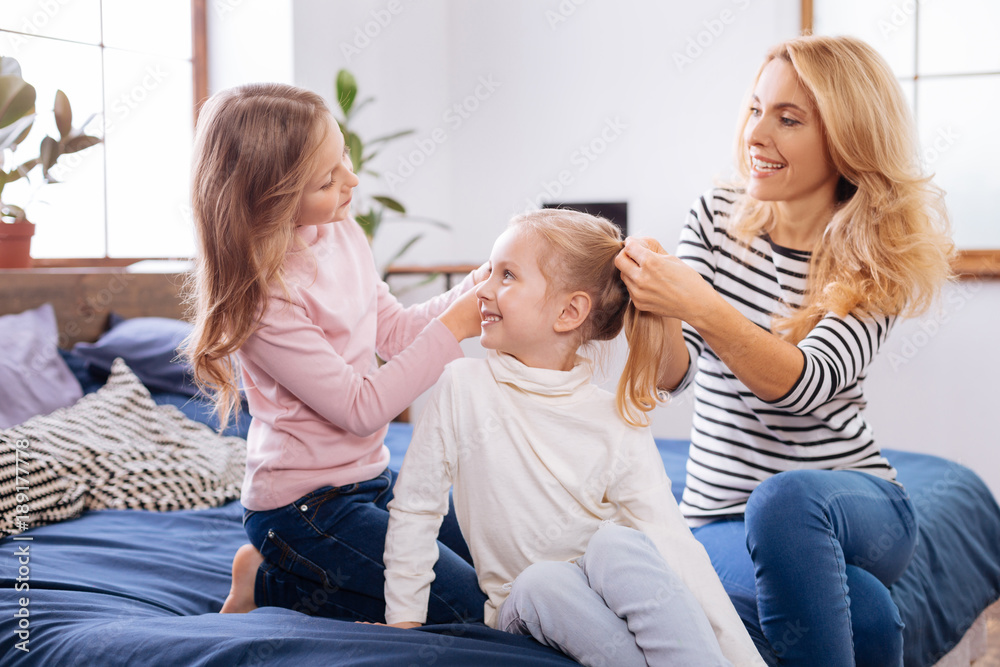 Family. Pretty joyful fair-haired girl smiling and making a hairdo for her younger sister together with her mom while sitting on the bed