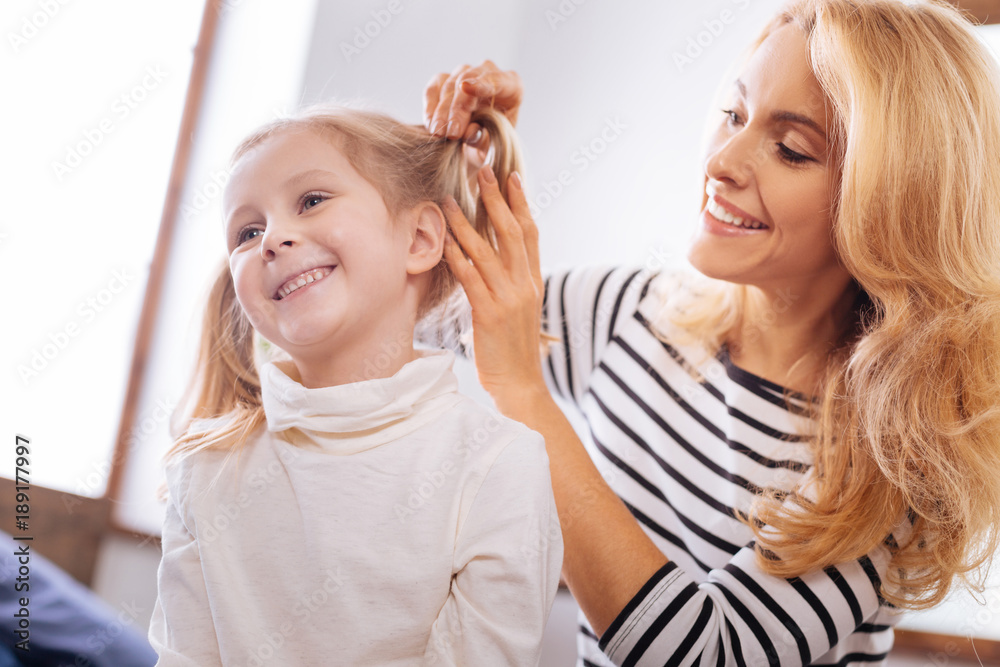 Happiness. Beautiful content blond young mother smiling and combing her daughters hair while sitting behind her child Happiness. Beautiful content blond young mother smiling and combing her daughters hair while sitting behind her child