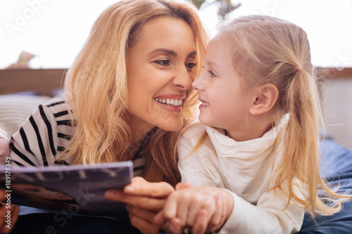 I cherish you. Loving joyful blond mother and her little daughter smiling and reading a magazine and mom caressing her