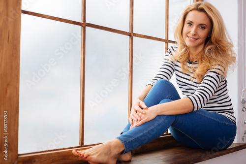 Relaxing. Attractive cheerful blond long-haired young woman smiling and wearing a shirt and jeans and sitting on the windowsill