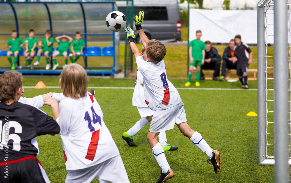 Obraz premium Young soccer goalkeeper save. Boy jumping and catching soccer ball. Football bench and children team in the background. Football match for children