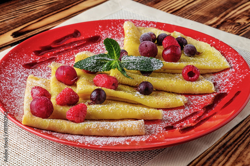 On the wooden table is a red plate. On it pancakes with berries and jam  sprinkled with powdered sugar and decorated with green mint leaves.