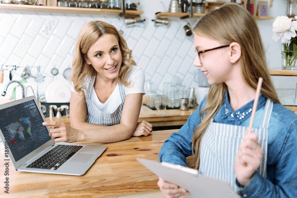 Look here. Attractive woman leaning on table and pointing at computer while standing opposite her daughter Look here. Attractive woman leaning on table and pointing at computer while standing opposite her daughter
