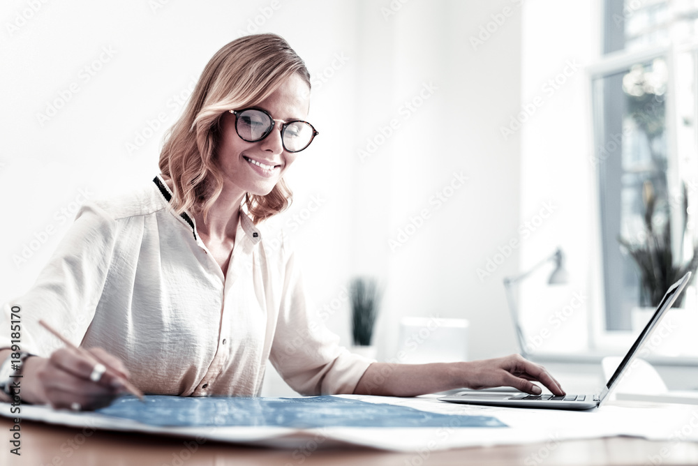At workplace. Pretty girl expressing positivity while looking at scheme, holding left hand on the keyboard of laptop At workplace. Pretty girl expressing positivity while looking at scheme, holding left hand on the keyboard of laptop