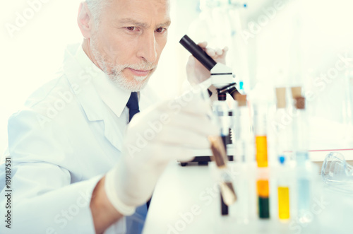 Let me have a look. Selective focus on a serious mature laboratory worker sitting at a microscope and taking one of the test tubes containing chemical liquid.