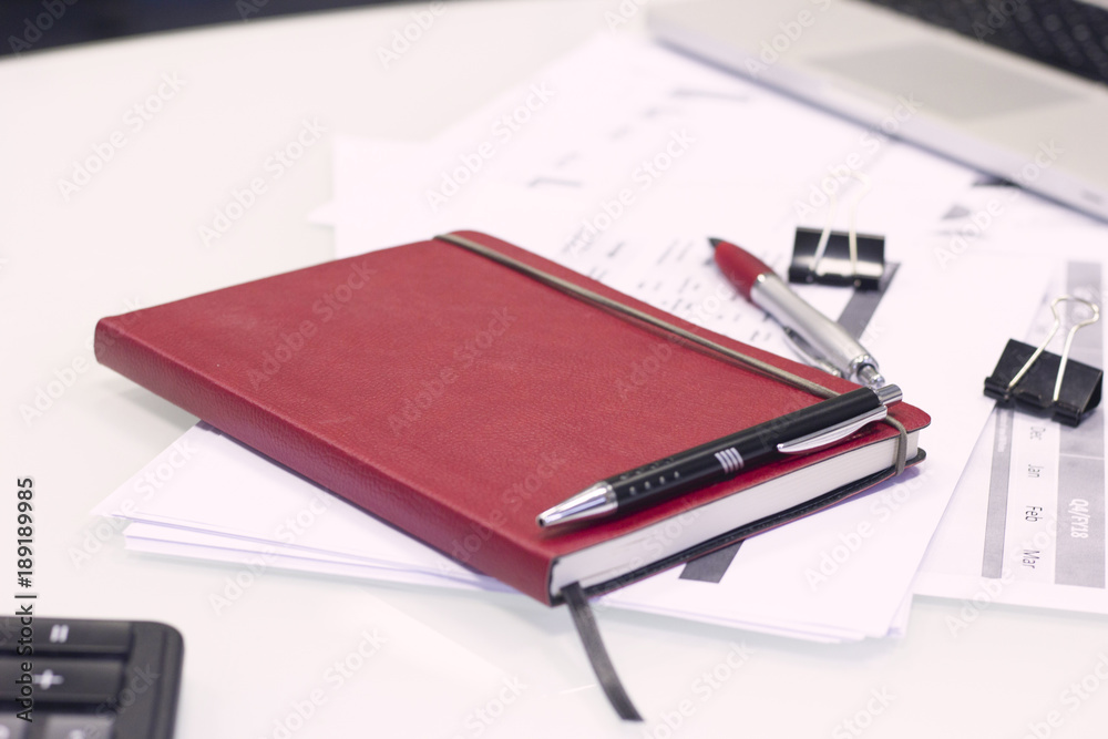 Business Office Workplace Concept . A planner in a red leather cover, black paper clips and a pen on printed graphs and charts, light background Business Office Workplace Concept . A planner in a red leather cover, black paper clips and a pen on printed graphs and charts, light background
