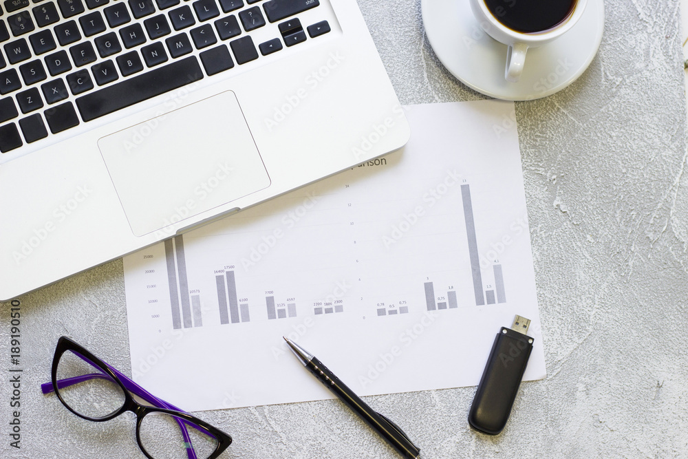 Modern office table desk, flat lay, top view. Workplace with a laptop, glasses, coffee cup, paper with graphs and USB flash drive, grey concrete background Modern office table desk, flat lay, top view. Workplace with a laptop, glasses, coffee cup, paper with graphs and USB flash drive, grey concrete background