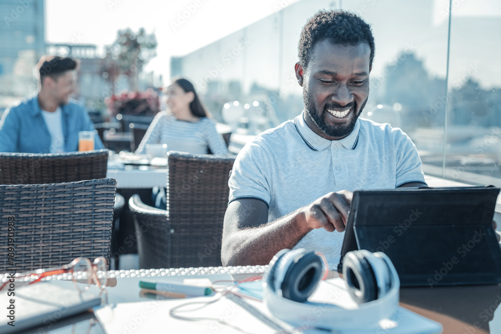 Browsing the Internet. Joyful young man smiling cheerfully while sitting in a local cafe and working on his digital tablet outdoors. Browsing the Internet. Joyful young man smiling cheerfully while sitting in a local cafe and working on his digital tablet outdoors.