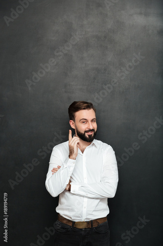 Brunette businessman posing on camera with tricky happy look, pointing index finger like he know something over dark gray background