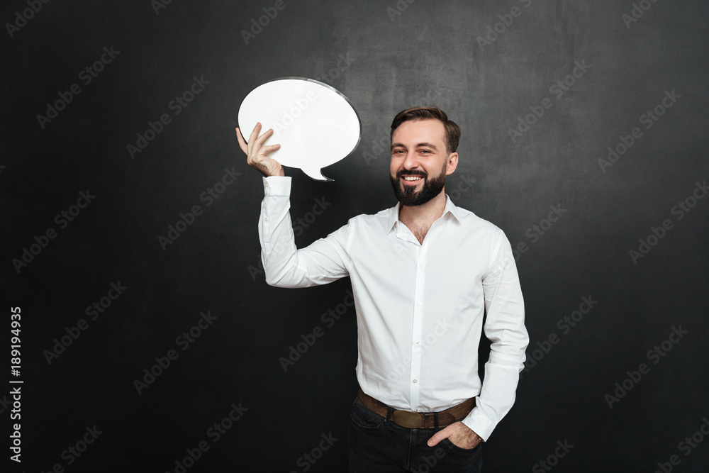 Good-looking brunette man holding blank speech bubble and looking at camera, over dark gray wall copy space