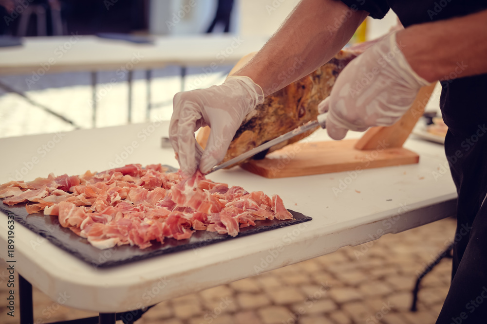 Closeup on chef hands slices prosciutto Italian delicatessen, restaurant party background. Festive time
