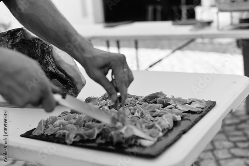 Closeup on chef hands slices prosciutto Italian delicatessen, restaurant party background. Festive time