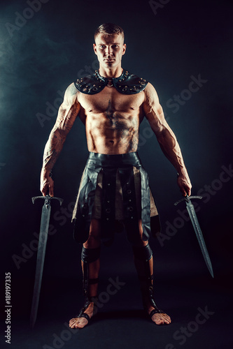 Portrait of handsome muscular gladiator with two swords. Studio shot. Black background. © zamuruev
