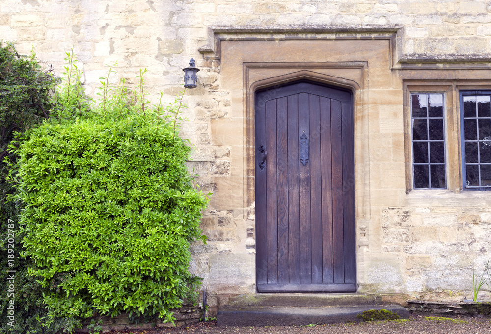 Dark brown wooden doors in an old traditional English lime stone house with evergreen shrubs on the side .