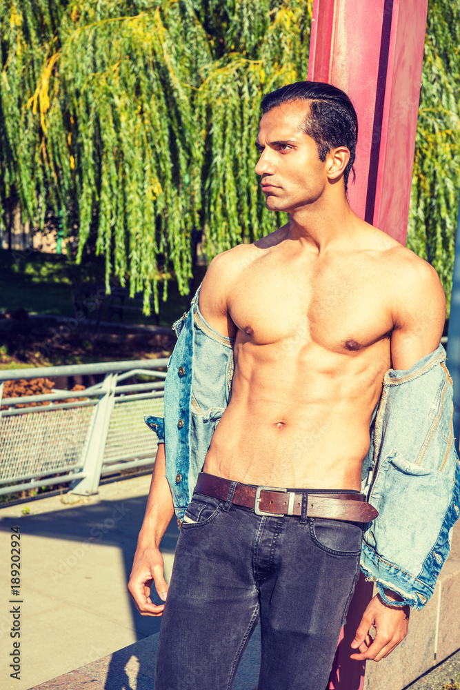 Young East Indian American Man wearing blue Denim jacket, opened, showing  half naked strong body, black jeans, standing against red pole at park in  New York, serious, looking up, thinking.. Stock Photo