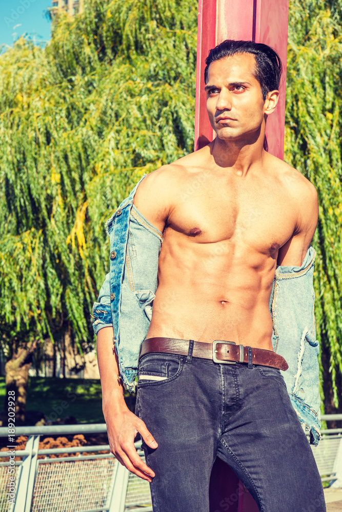 Young East Indian American Man wearing blue Denim jacket, opened, showing  half naked strong body, black jeans, standing against red pole at park in  New York, serious, looking up, thinking.. Stock Photo