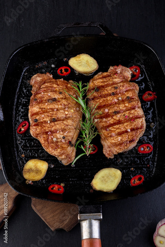 Grilled pork steak in grill pan with rosemary, pepper chilli and ginger on wooden board.