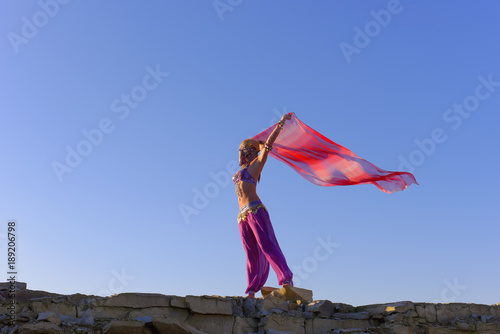 A beautiful blonde oriental belly dancer dressed in a purple  colored belly dance costume relaxes in a large stone quarry. She spends her leisure time in and around the large stones relaxing.