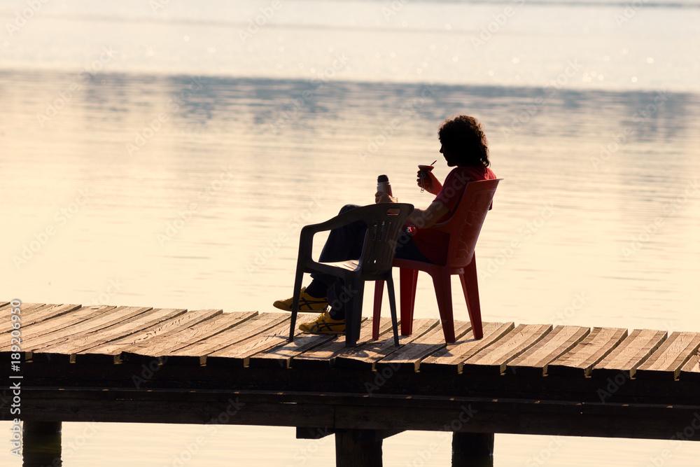 A young man sitting on the pier, relaxes enjoying a drink by the lake, in the light of the sunset.