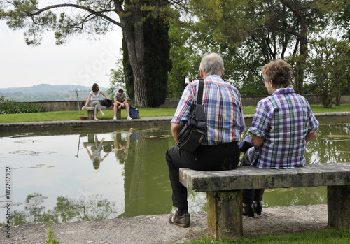 Romantic old couple sitting on park bench by lake. In front of them there is a couple of young lovers who fight.