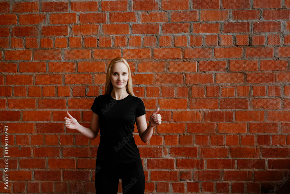 Portrait of beautiful blonde woman in black sportswear standing against the background of a red brick wall after fitness workout.Fites concept. The girl poses, the place for copy spase nearby Portrait of beautiful blonde woman in black sportswear standing against the background of a red brick wall after fitness workout.Fites concept. The girl poses, the place for copy spase nearby