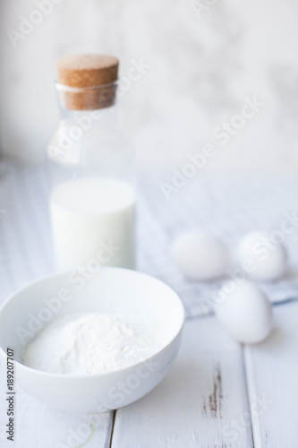 Baking Homemade Pastry Concept. A bowl of flour, eggs and a glass bottle of milk with cork. White wood and white plaid napkin as a background