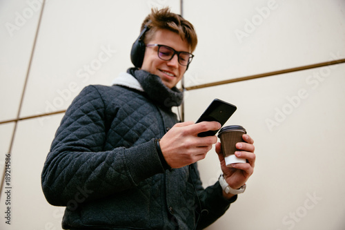 Smiling handsome man in eyeglasses listening to music in headphones, while looking at mobile screen, holds cup of coffee, dressed in warm jacket and jeans. Outdoors.