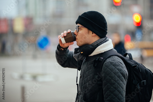 Attractive guy in eyeglasses drinking coffee, outdoors. Dressed in warm jacket and hat, with headphones and school bag. Side view.