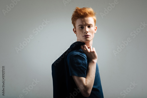 Look. Handsome humourless red-haired young man carrying a black jacket on his shoulder and thinking and looking over his shoulder