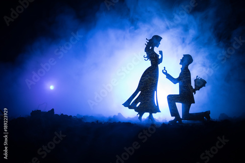 Will you marry me? Silhouette of young man staying on the knee and making proposal for his lovely girl on the beach against dark toned background. Valentine greeting card decor