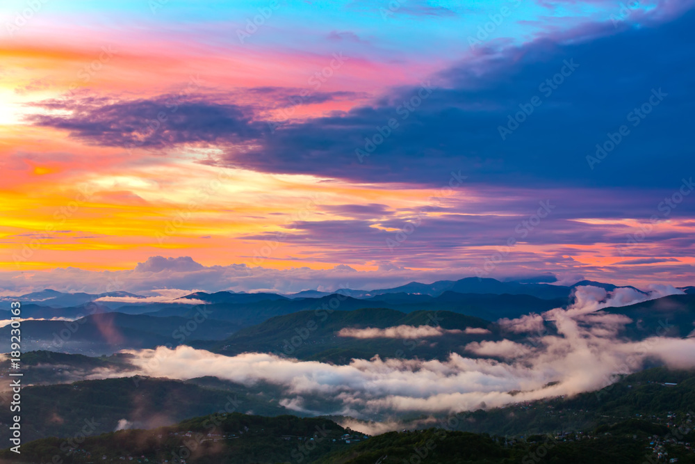 The mountain valley in the evening in the fog is lit by the sunset under beautiful colorful clouds of yellow blue violet red. Panorama of the coast from the observation tower on Mount Akhun.