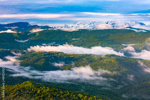 The valley is covered with a spring green forest with rare clouds and fog on the back of the majestic great snowy peaks. Panorama of the Caucasian mountains from the observation tower on Mount Akhun.