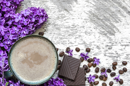 Cup of coffee with coffee beans  chocolate and lilac flowers over white wooden background