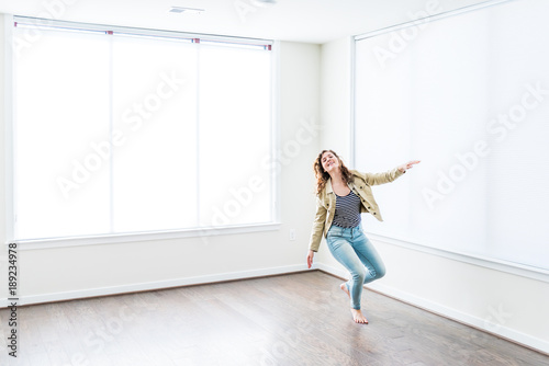Fototapeta Naklejka Na Ścianę i Meble -  One young happy smiling woman jumping up in empty modern new room with hardwood floors and large sunny windows in apartment
