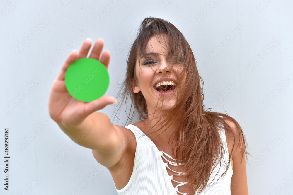 A young girl stands in front of a grey cement wall. She holds up a round green card to the camera. She shows a positive expression on her face. Green for go is the message she is sending. A young girl stands in front of a grey cement wall. She holds up a round green card to the camera. She shows a positive expression on her face. Green for go is the message she is sending.
