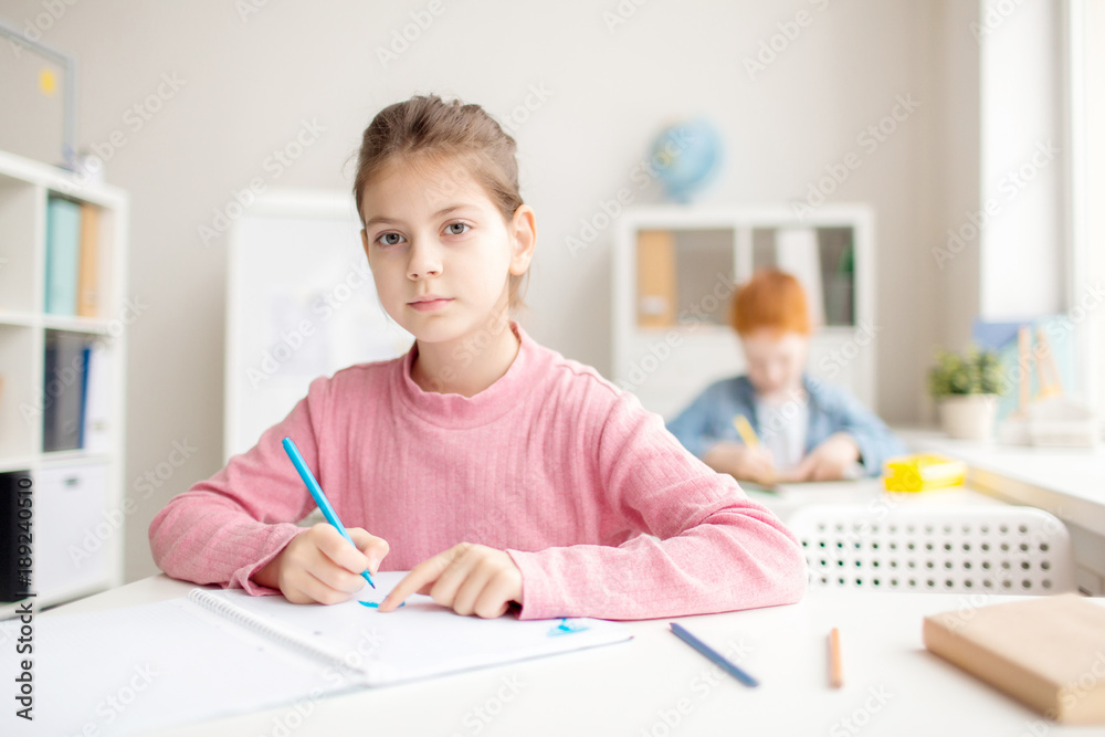 Little schoolgirl wih blue highlighter drawing by workplace with classmate on background Little schoolgirl wih blue highlighter drawing by workplace with classmate on background