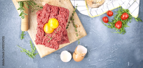 Fresh beef meat in forcemeat, on a wooden board. Raw eggs and tomatoes are small. Herbs for cooking and round buns. On a gray stone background. Free space for writing text.