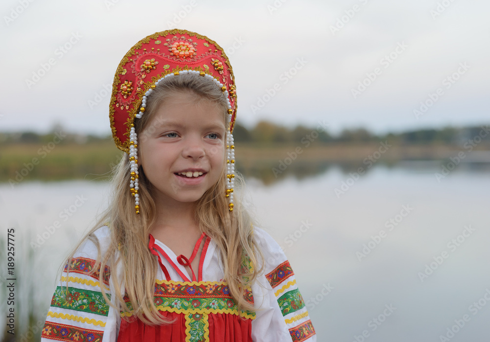 Portrait of smiling little Caucasian blond girl in Russian national costume on blur natural landscape background in summer