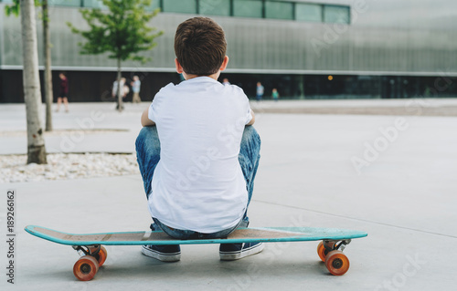Back view. The boy, dressed in a white T-shirt and blue jeans, sits on a city street on a skateboard. On background modern building in soft focus. Lifestyle. Space for text, logo, image. Mock up.