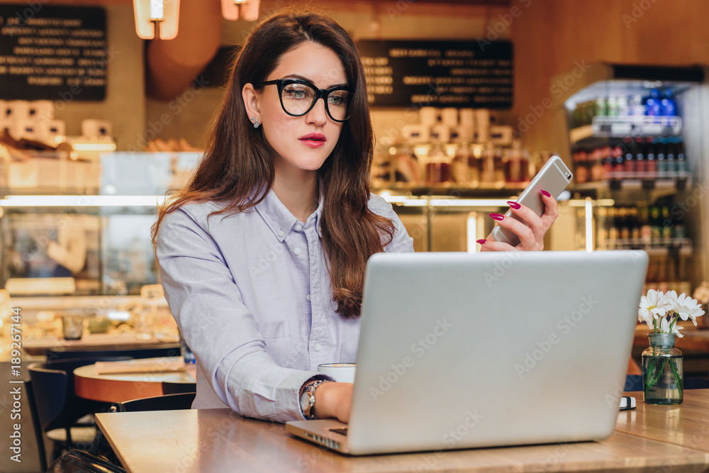 Front view. Young businesswoman is sitting in cafe at table, working on laptop. Hipster girl blogging, browsing internet, learning online, checking email. Online marketing, education, e-learning. Front view. Young businesswoman is sitting in cafe at table, working on laptop. Hipster girl blogging, browsing internet, learning online, checking email. Online marketing, education, e-learning.