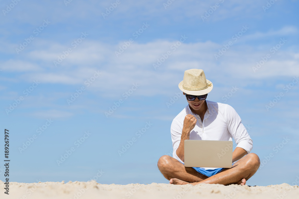 Asian traveller man using laptop on the beach with blue sky background. Man is success pose.