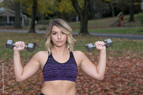 Sexy Caucasian female fitness model lifts weights outdoors in fall in purple sports bra and black leggings