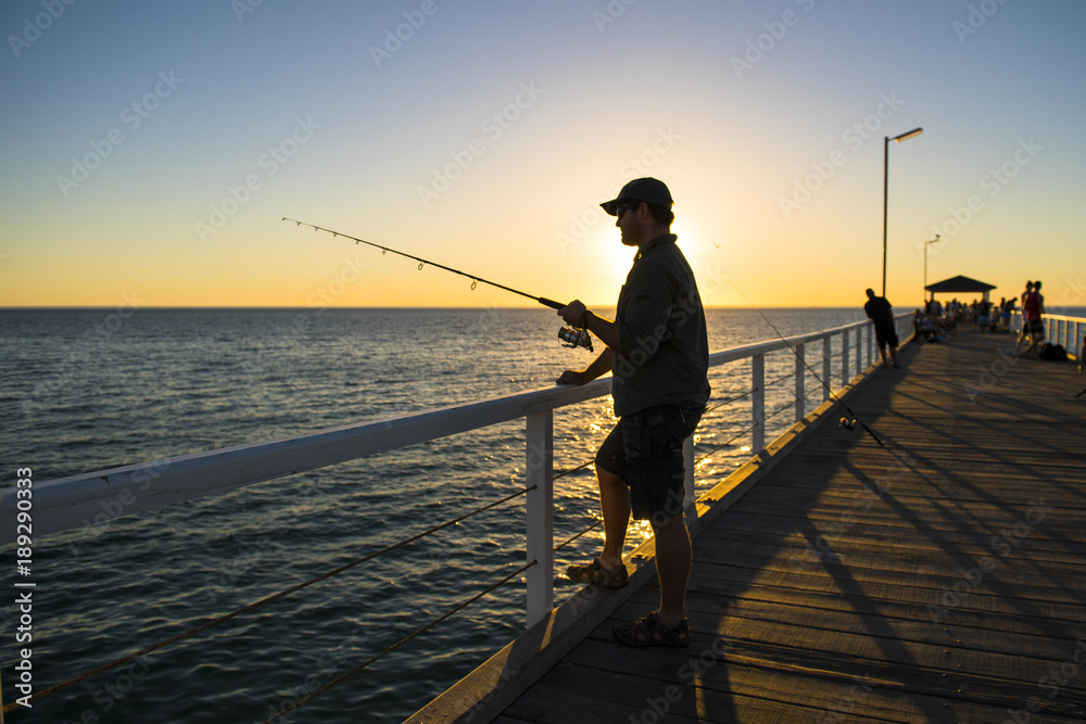silhouette of fisherman with hat and fish rod standing on sea dock fishing at sunset with beautiful orange sky in vacations relax hobby