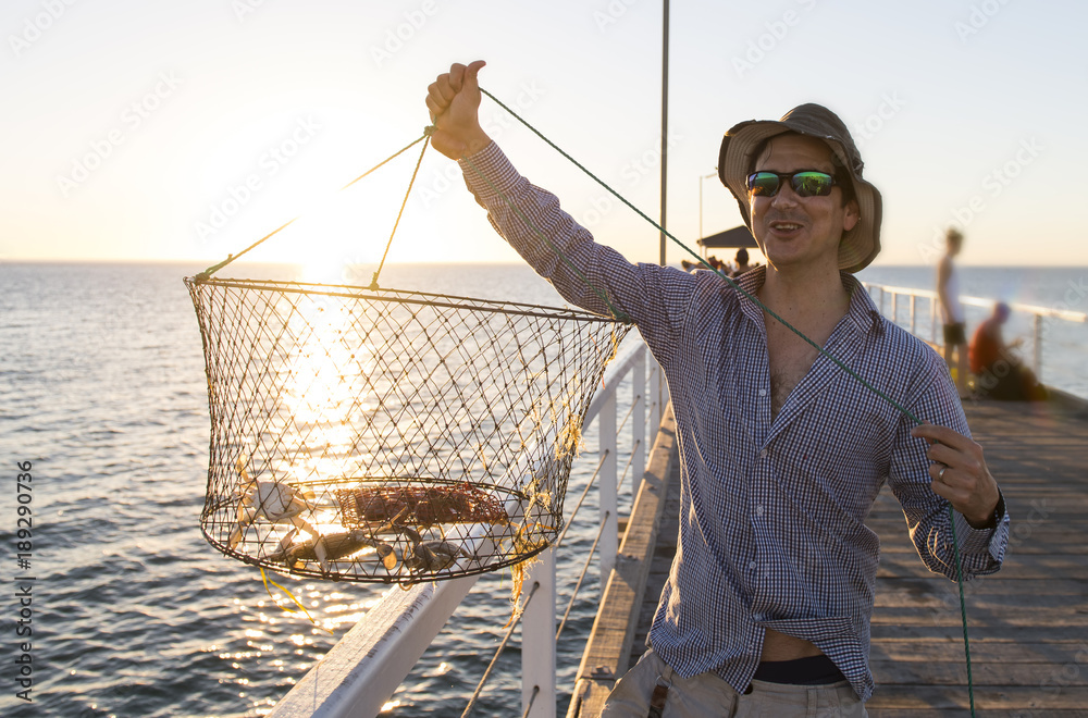 proud attractive fisherman showing fish and crabs basket net captures  smiling at sea dock sunset in man fishing as weekend hobby Stock Photo